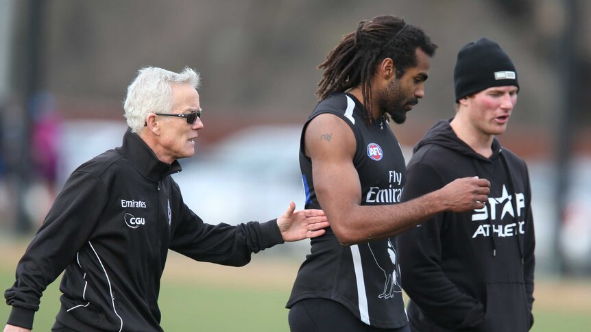 Heritier Lumumba walks at Collingwood AFL training with two members of the Magpies staff.