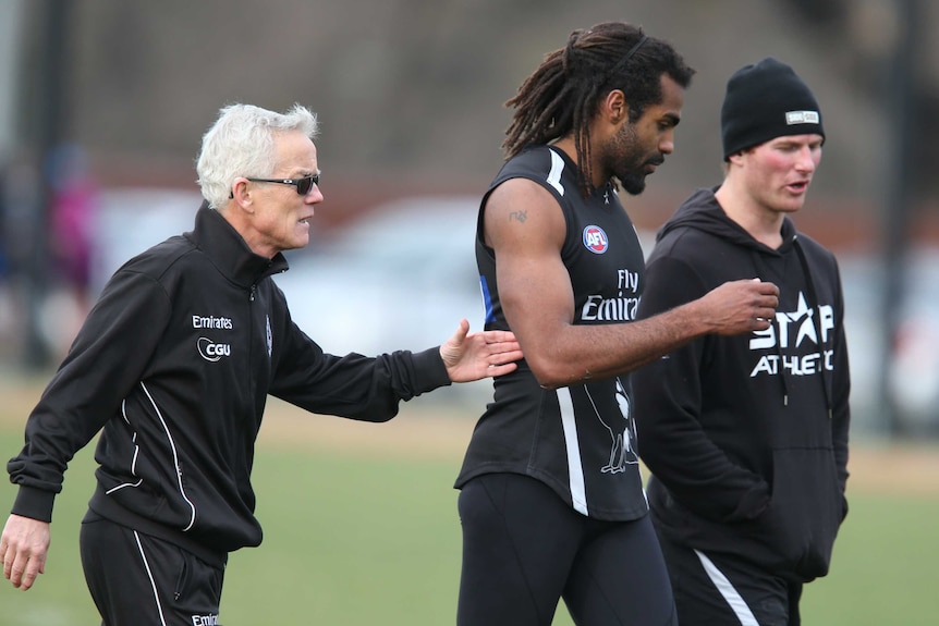 Héritier Lumumba walks at Collingwood AFL training with two members of the Magpies staff.