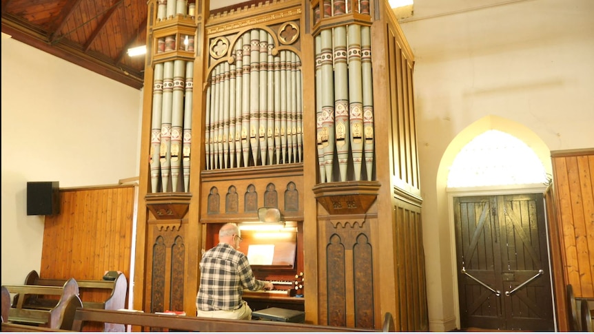 A man sits and plays a huge wooden and grey steel piped organ that towers over him in a small church