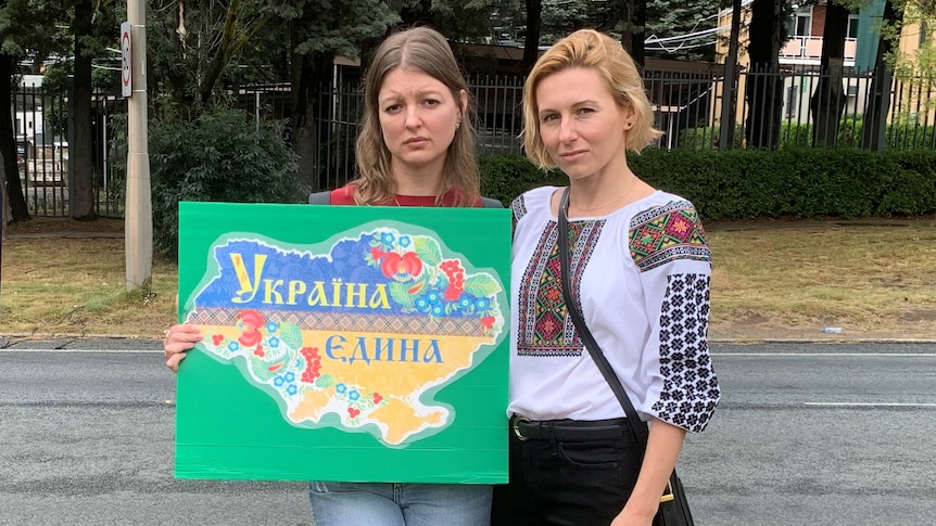Two woman stand holding a sign which reads 'Ukraine One' in Cyrillic text.