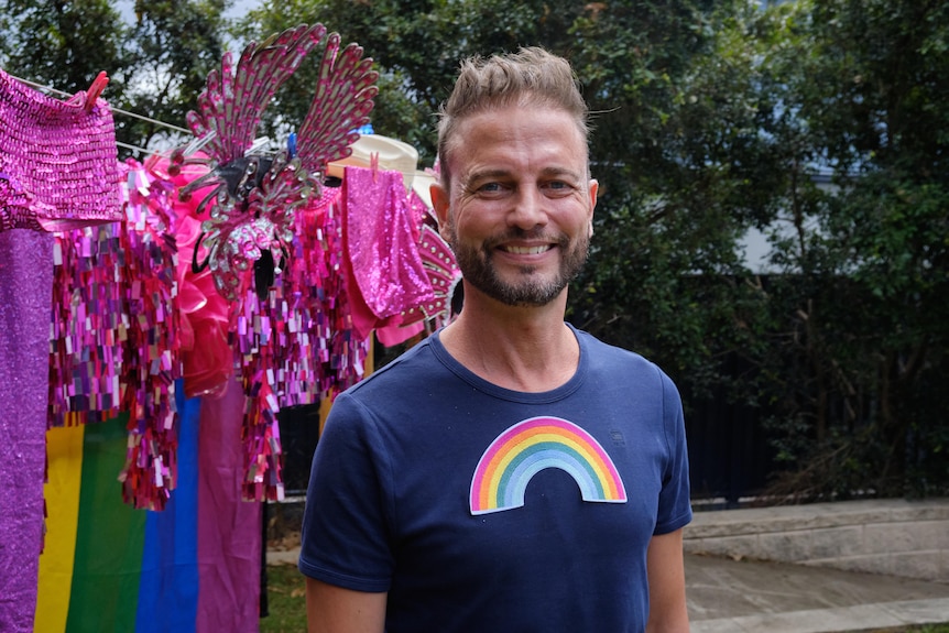 Man wearing blue t-shirt in backyard, with rainbow-decorated clothes line in background. 