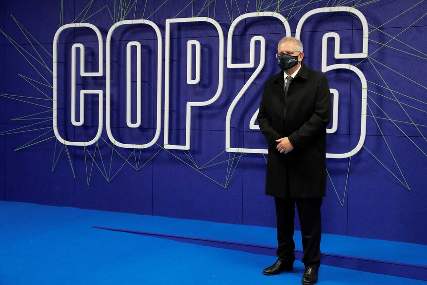 man in suit and mask standing in front of  lit up COP26 sign