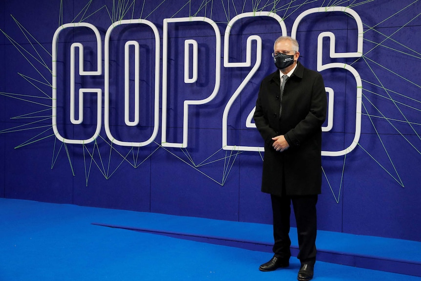 man in suit and mask standing in front of  lit up COP26 sign