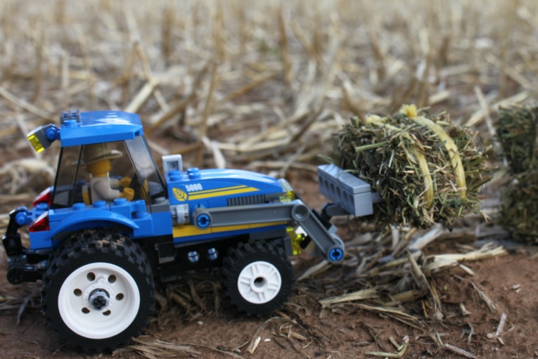 Close up on a LEGO farmer tractor picking up a mini hay bale in a ploughed field.