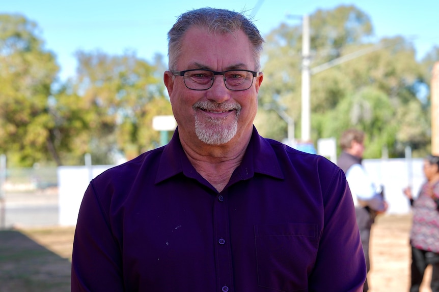 A white man with glasses and a maroon collared shirt smiling on a sunny day. 