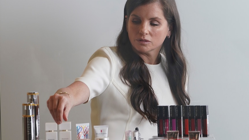 A well dressed middle-aged woman extends a hand towards numerous make-up products on display