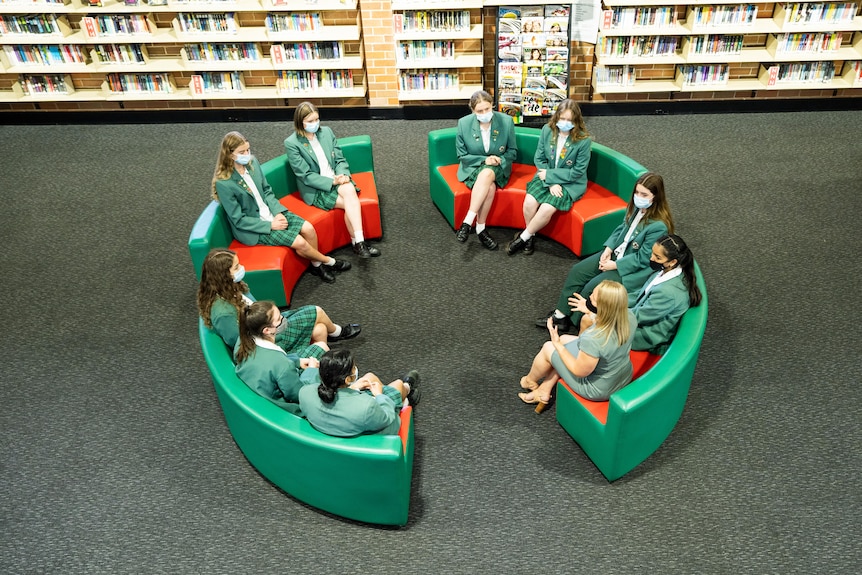 Nine schoolgirls in green formal jackets and one teacher sit on couches in a circle, in a large library space.