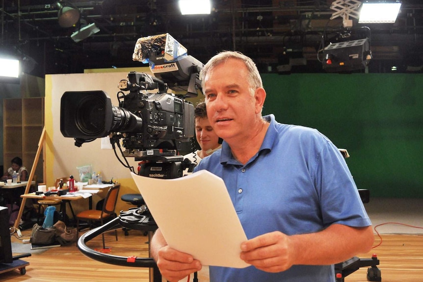 TV producer Ian Munro stands beside a TV camera and operator while directing in a studio.