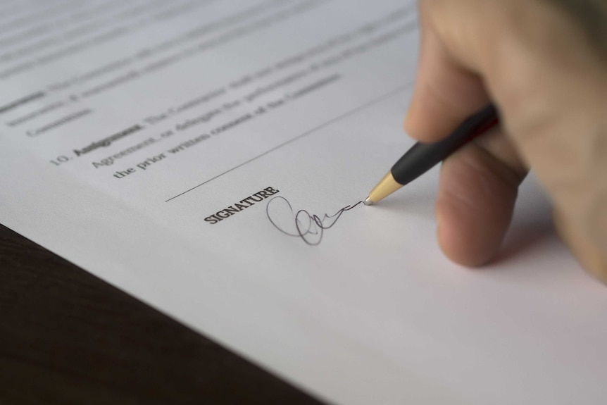 A hand signs a signature with a pen at the end of a form or contract.