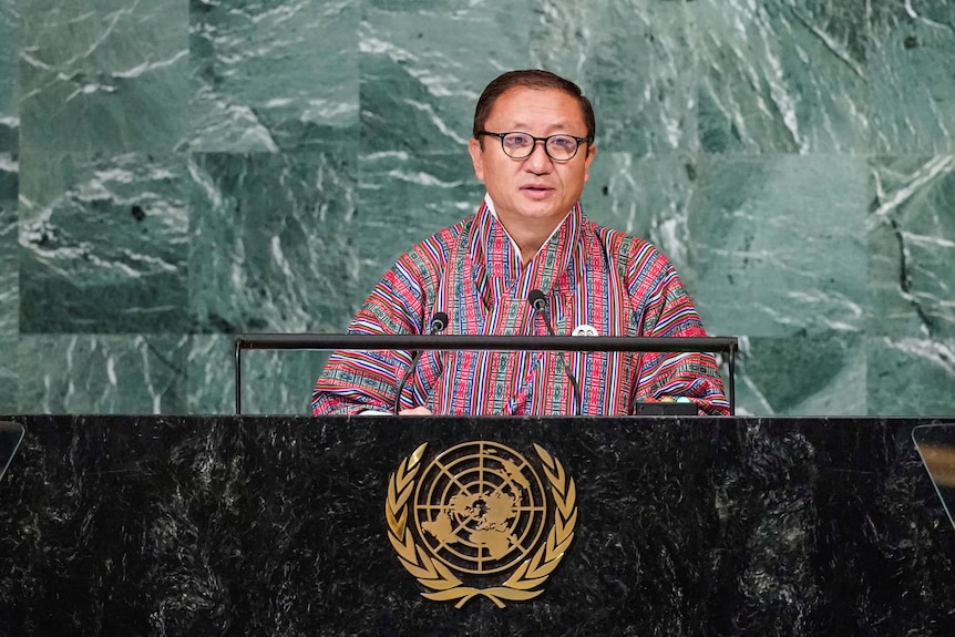 Un butanés con anteojos vestido con ropa tradicional roja y azul se para detrás del podio principal de la Asamblea General de las Naciones Unidas.