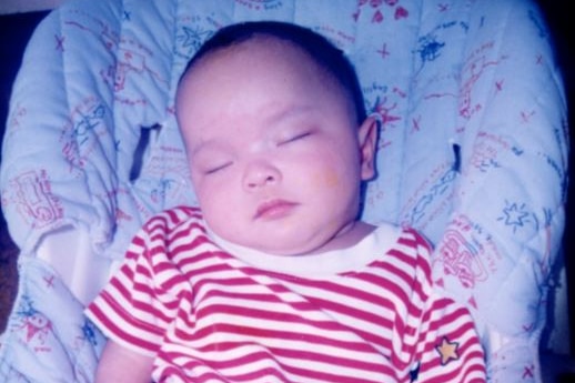 A small Asian baby is seen appearing to sleep in a rocker. He has a head of dark hair and wears a red and white striped shirt.
