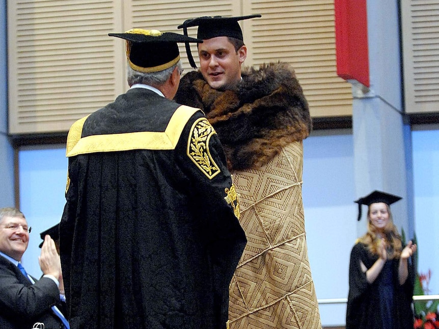 A young man in a graduation cap and possum skin cloak shakes hands with a university chancellor