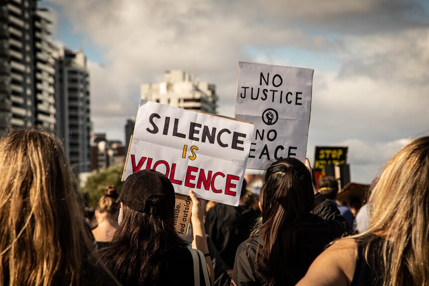 A group of people at a rally holdings signs that say 'silence is violence' and 'no justice, no peace'.