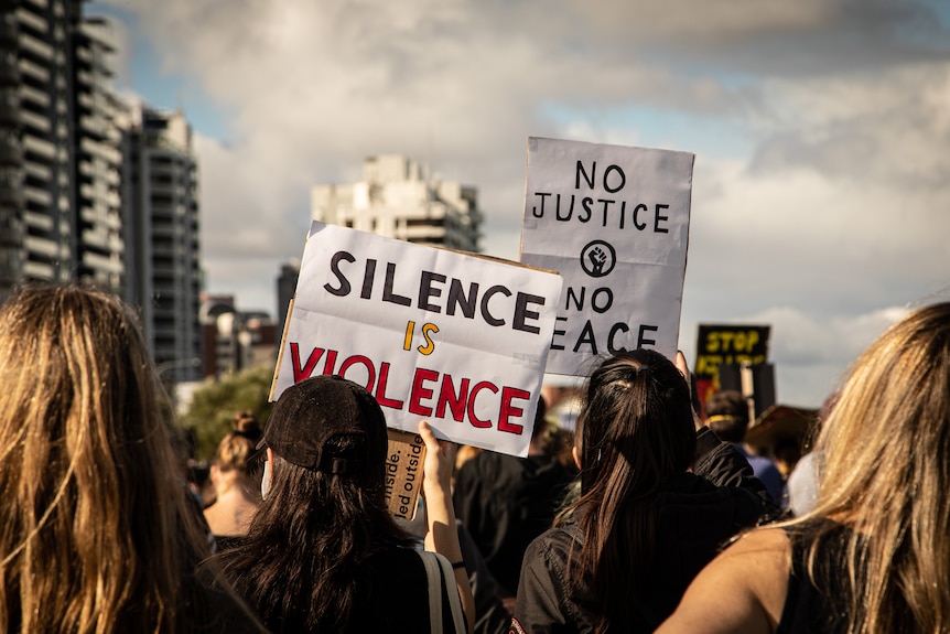 A group of people at a rally holdings signs that say 'silence is violence' and 'no justice, no peace'.