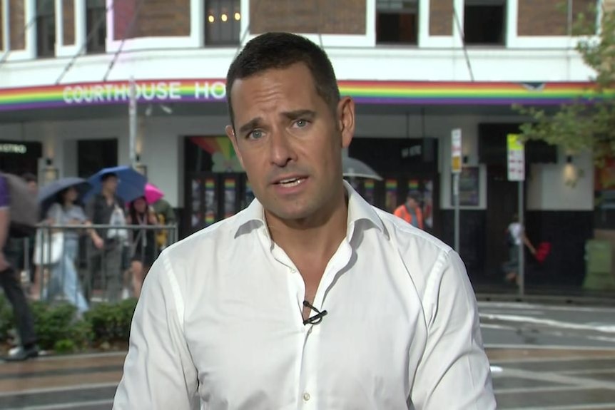 A man in a white shirt stands giving a speech, standing in front of a pub with a rainbow signage