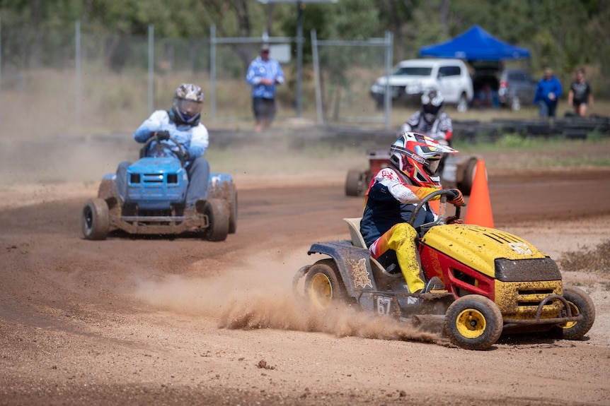 Three people wearing motorbike helmets race ride-on lawn mowers around a dirt track.