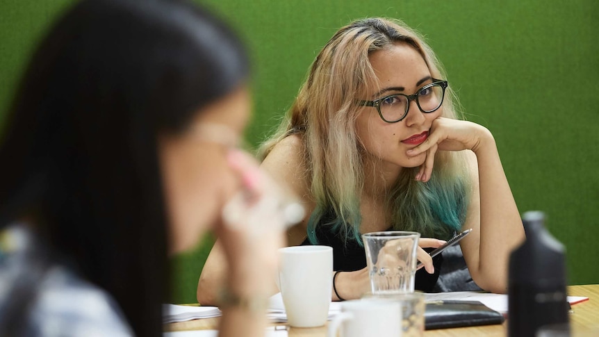 Seated behind an out-of-focus woman in the foreground, Anchuli Felicia King rests her elbow on a desk.