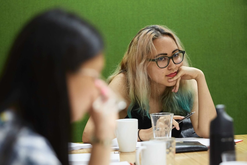 Seated behind an out-of-focus woman in the foreground, Anchuli Felicia King rests her elbow on a desk.