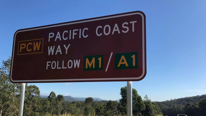 A large brown sign with white lettering that reads Pacific Coast Way next to a double lane bitumen road with a car driving past