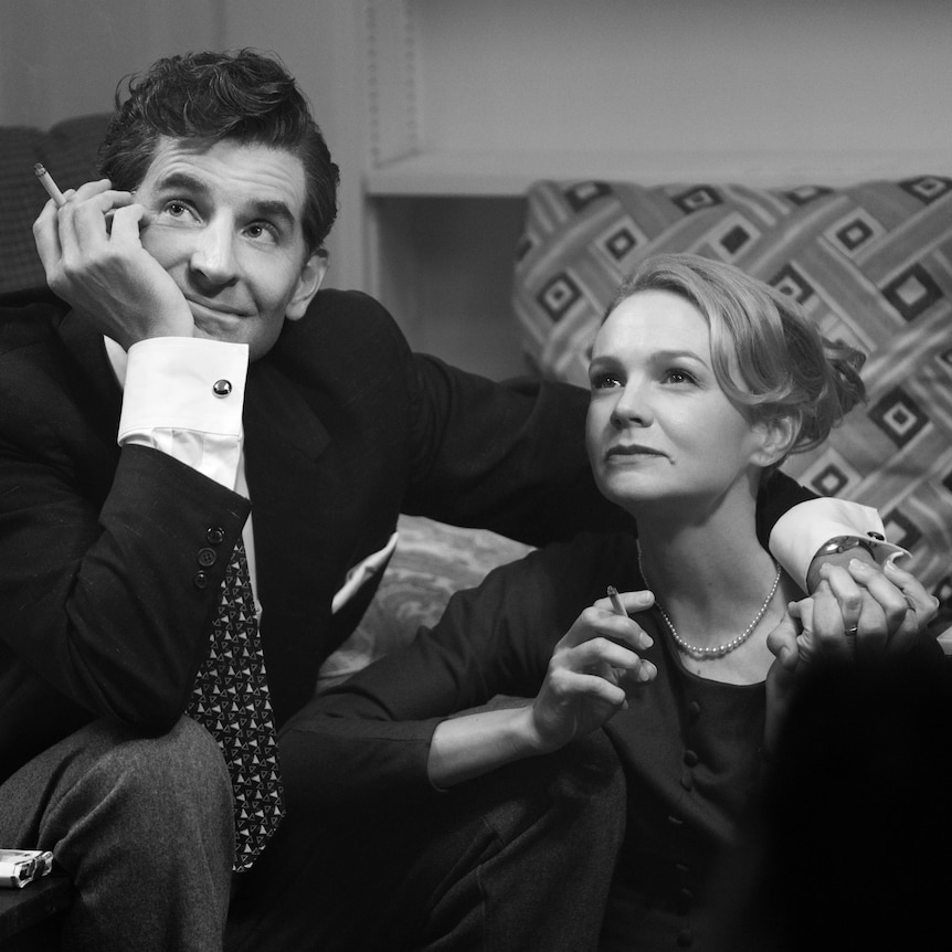 A black and white still of Bradley Cooper and Carey Mulligan seated and holding hands as Bernstein and his wife.