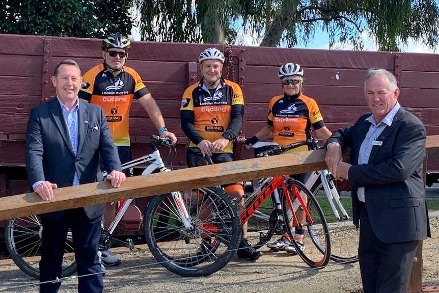 Two men wearing suits and three cyclists wearing cycling gear with bikes standing in front of heritage train freight carriage.