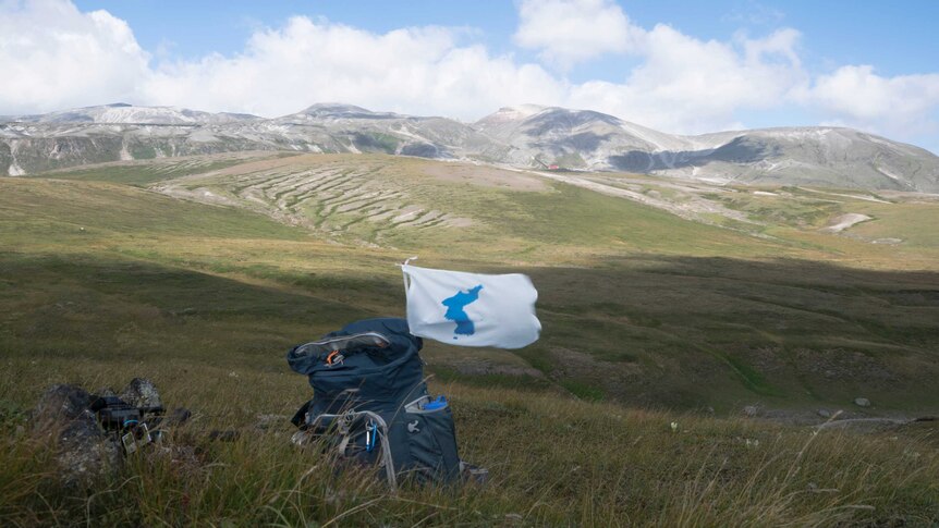 A Korean Unification flag is attached to a backpack placed on a field with mountains in the background.