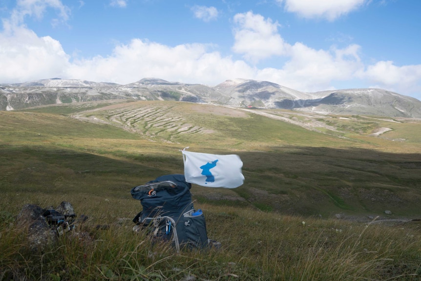 A Korean Unification flag is attached to a backpack placed on a field with mountains in the background.