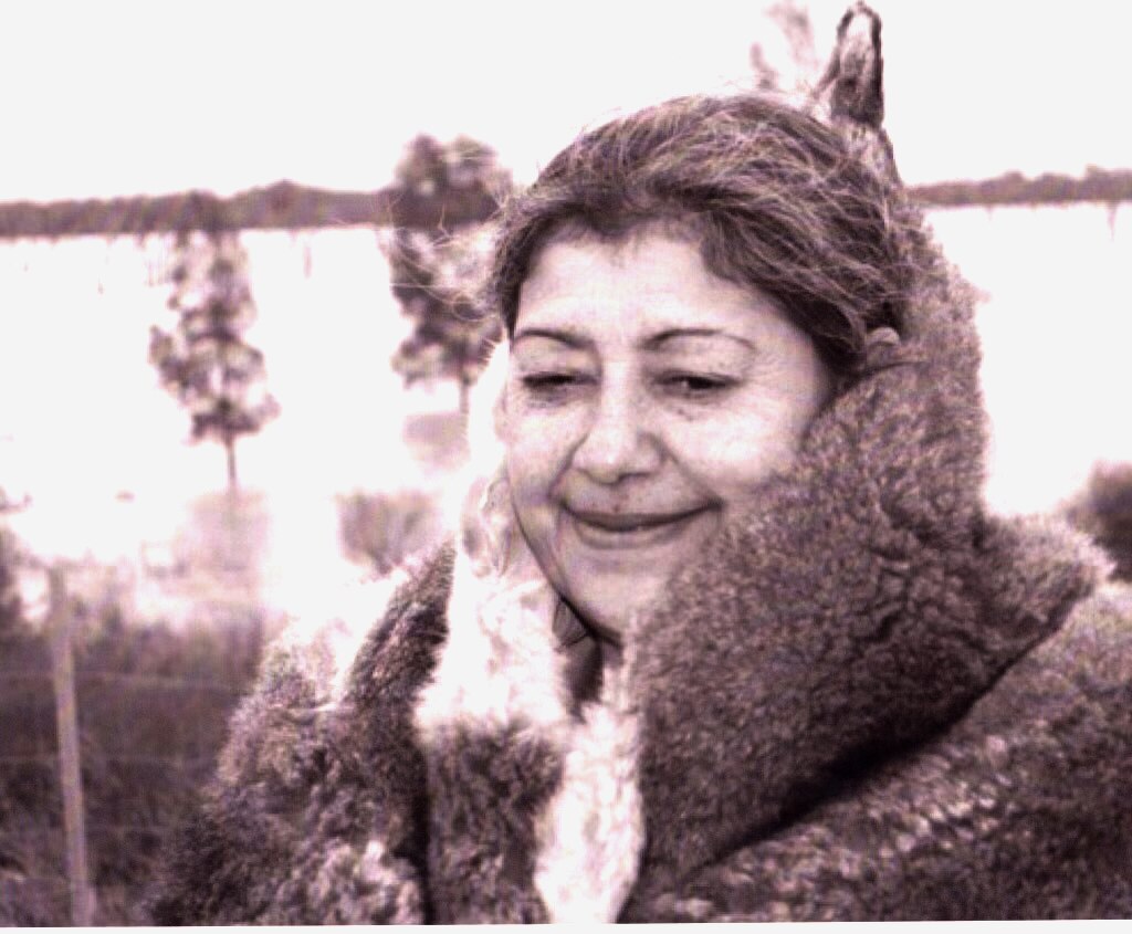 Aunty Margaret Gardiner smiles, wearing a possum-skin cloak as she stands in front of a field in a black-and-white photo.