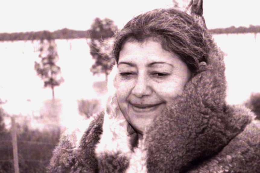 Aunty Margaret Gardiner smiles, wearing a possum-skin cloak as she stands in front of a field in a black-and-white photo.