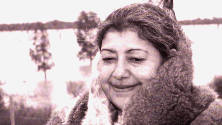 Aunty Margaret Gardiner smiles, wearing a possum-skin cloak as she stands in front of a field in a black-and-white photo.