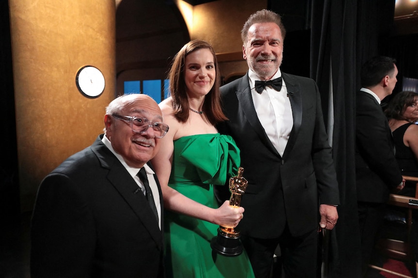 Three people pose in formal wear, with a woman in green holding an Academy Award 