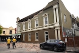 A two-storey building crumbles from the top as two construction workers walk by. 