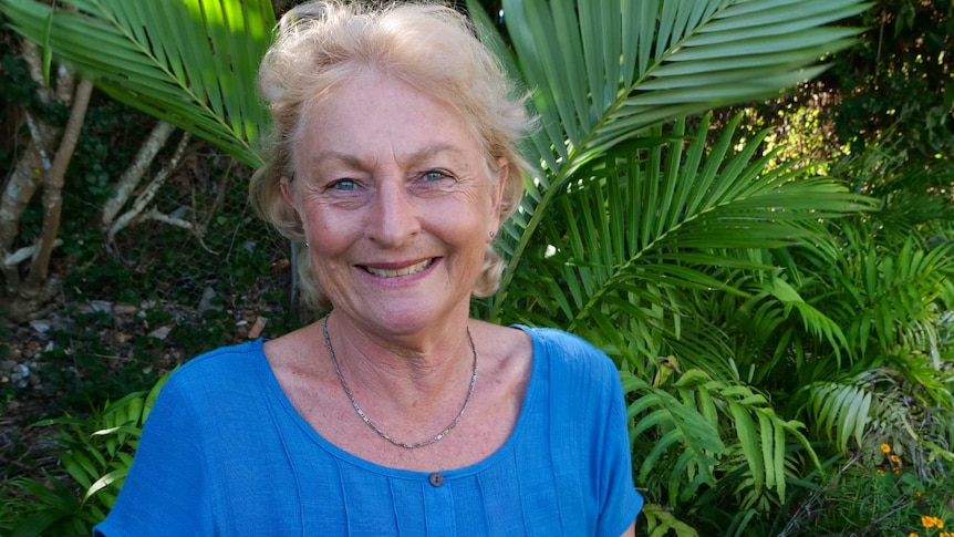 Lynne Barrett wearing a blue shirt sits in a garden with her hands in her lap, smiling, blue eyes and short blonde hair.