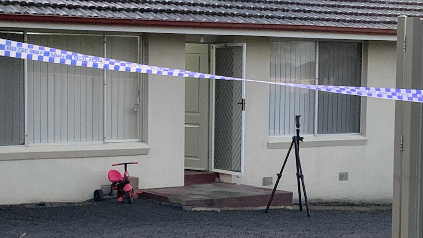 Police tape cordons off the driveway to the house, where a pink trike sits near the front door.