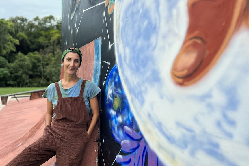 A woman stands smiling at the camera beside a concrete wall at a skate park with a mural spray painted on it.