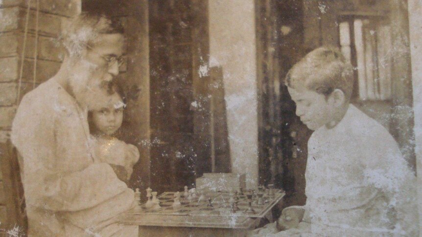 A man sits at a table with his children, playing chess in a sepia-toned image.