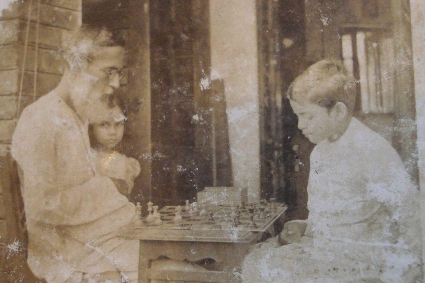 A man sits at a table with his children, playing chess in a sepia-toned image.