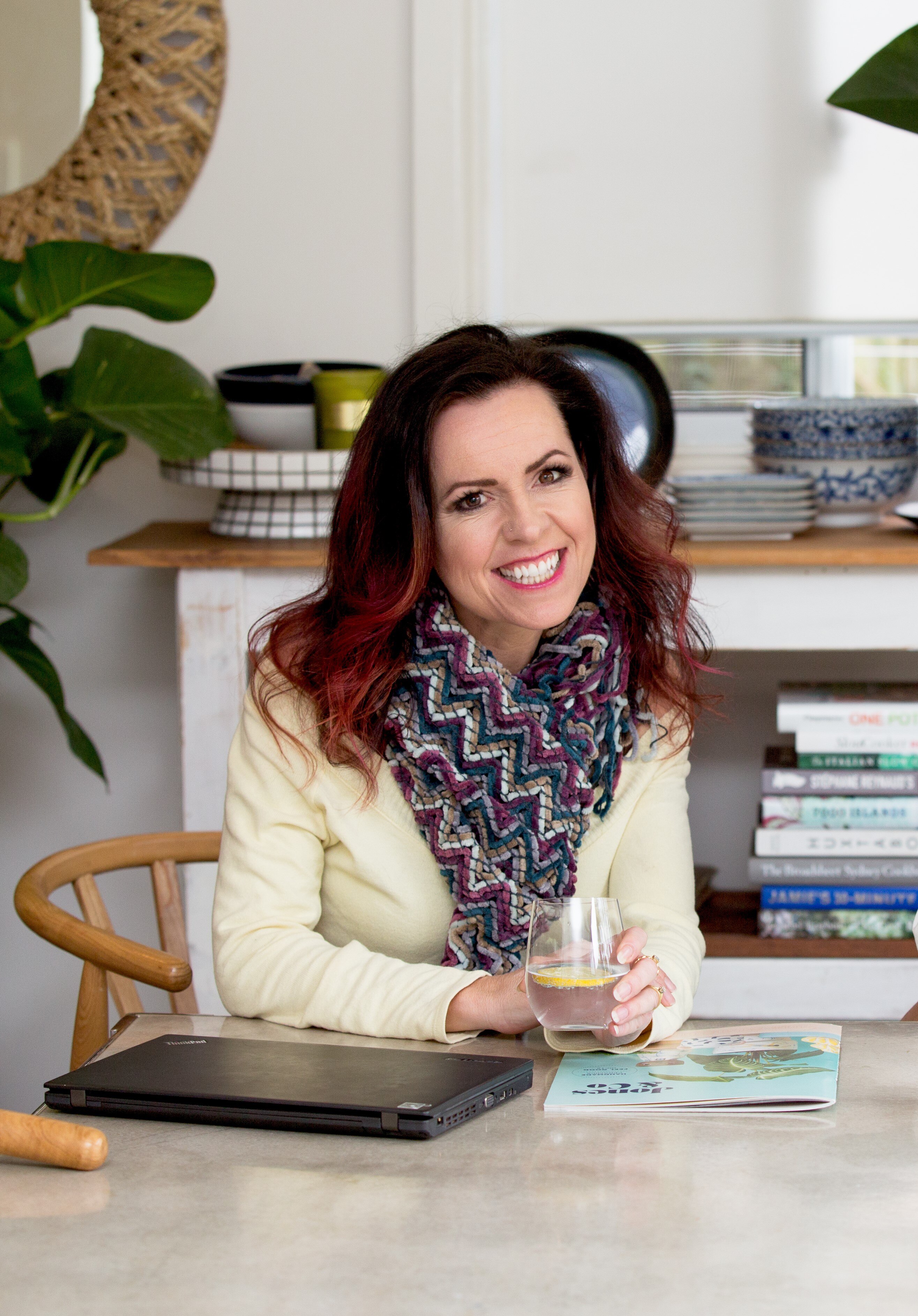 Nicole is wearing a light yellow jacket with a scarf sitting at a table with a glass of water in her hand and smiling