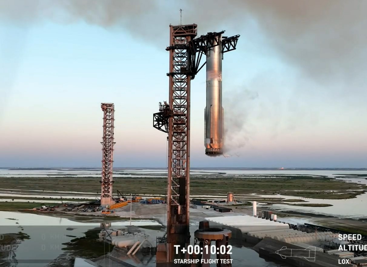 Smoke is emitted from a large silver cylinder as it hangs from two large metal prongs on a launch tower.