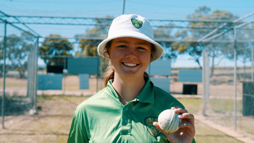 A young woman with fair skin and brown hair smiles at the camera holding a cricket ball wearing a bucket hat