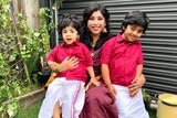 Preethi smiles in her backyard, holding her two young sons who wear matching red shirts.