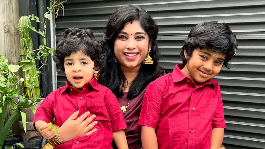 Preethi smiles in her backyard, holding her two young sons who wear matching red shirts.