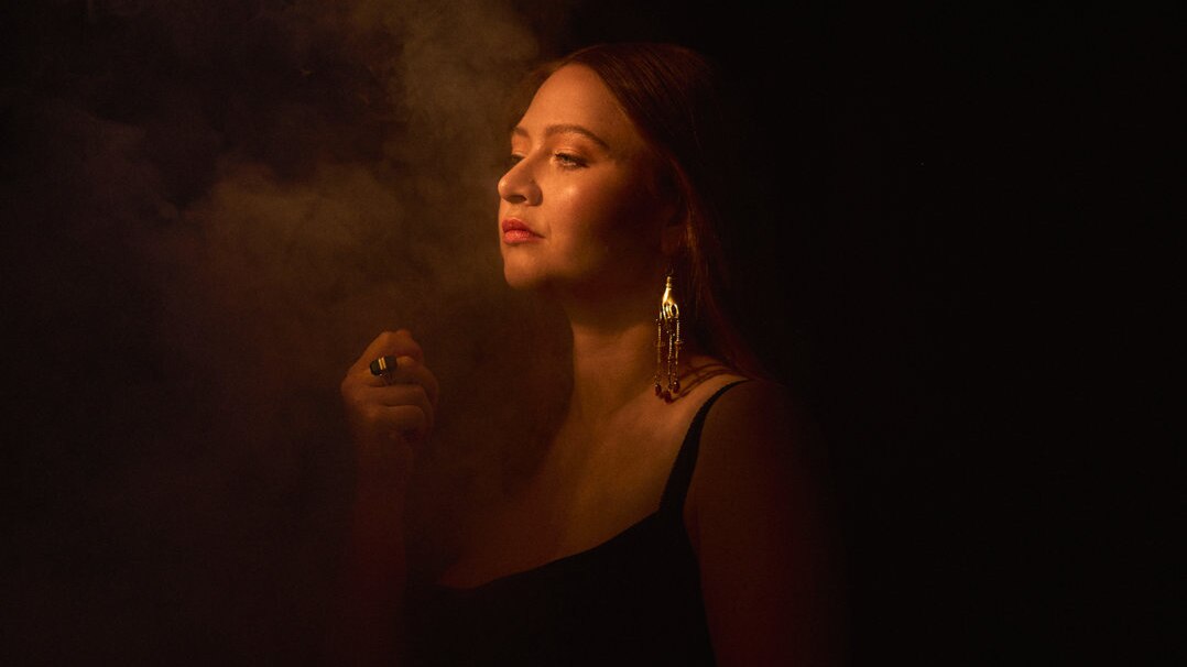 Trumpeter and singer AUdrey Powne surrounded by fog, taken in a dark space; she's lit by a warm, yellow light