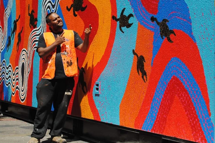 Gayiri man Yamba Konrad Ross stands in front of a wall of Aboriginal art.