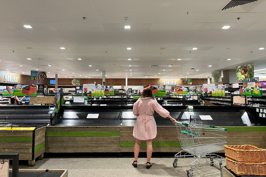 A woman holding a trolley and looking at empty shelves in the fresh produced section of a Woolworths supermarket.