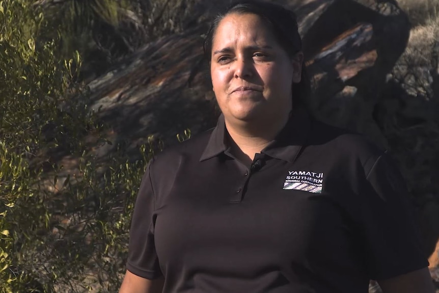 An Aboriginal woman standing in front of a rocky ridge in the bush. 