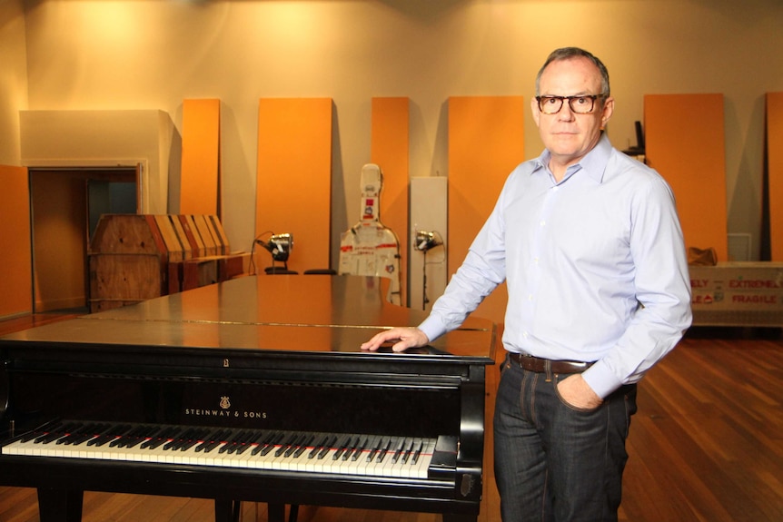 A man in glasses and a business shirt next to a piano