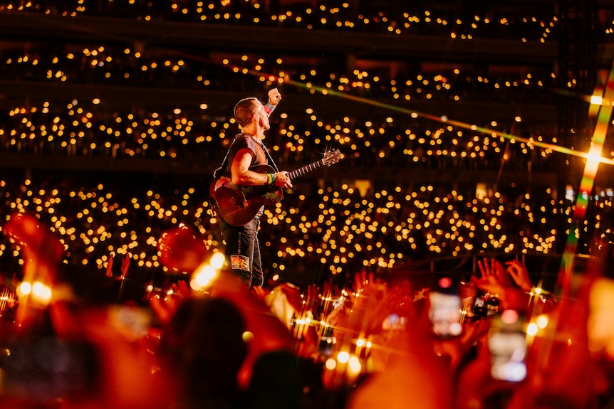 Chris Martin on stage in Perth surrounded by audience members holding yellow lights.