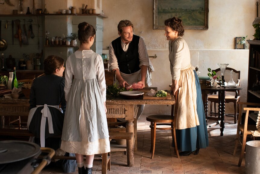 A film still of Benoît Magimel and Juliette Binoche, in late C19 dress, standing together around a table, with two women.