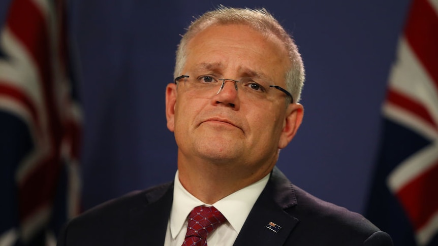 Prime Minister Scott Morrison in business attire at press conference in foreground, with Australian flags blurred in background.